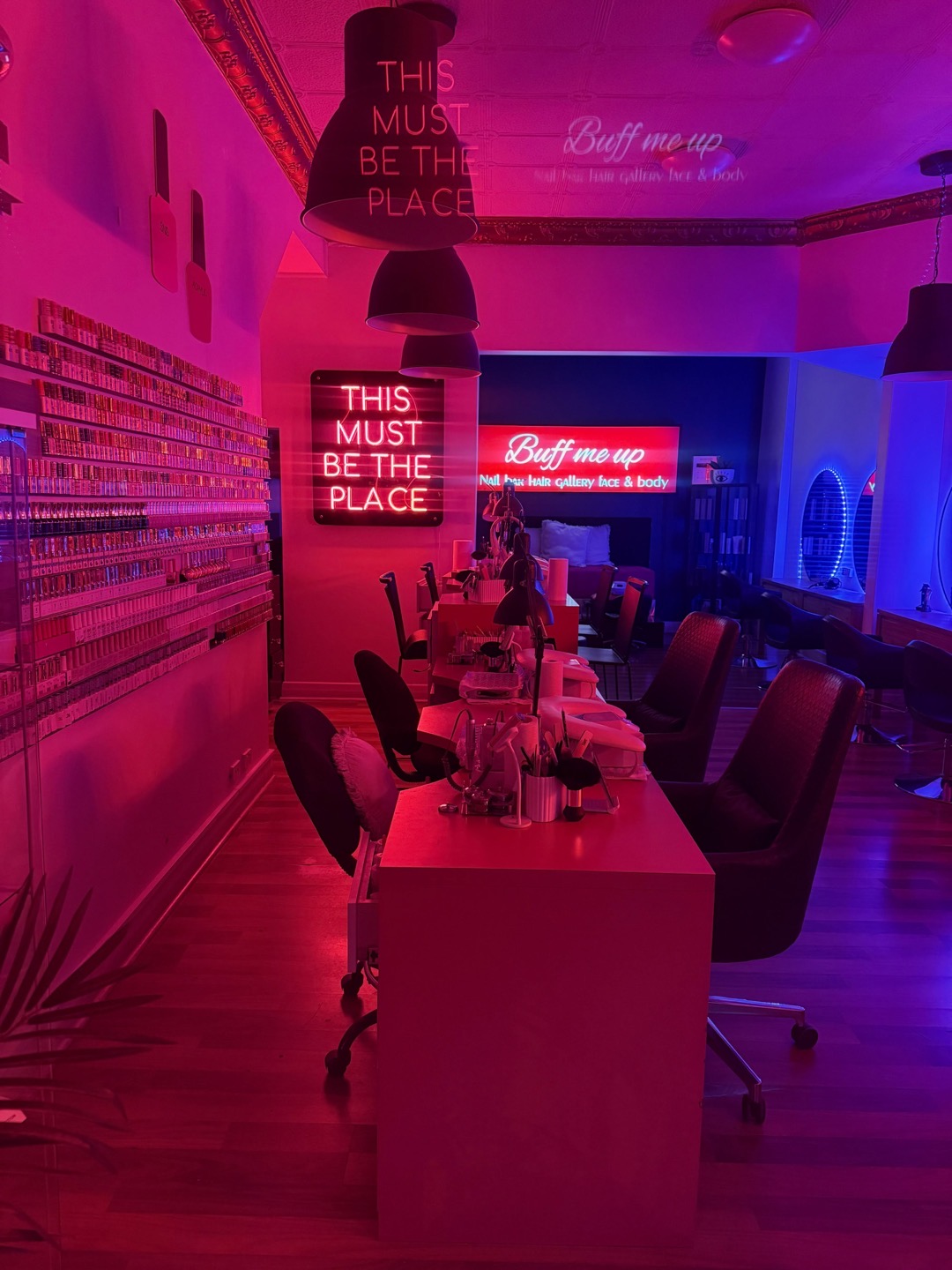 Inside Buff Me Up at night: nail desks under pink light, a full wall of polish, and neon signs reading This Must Be The Place and Buff Me Up
